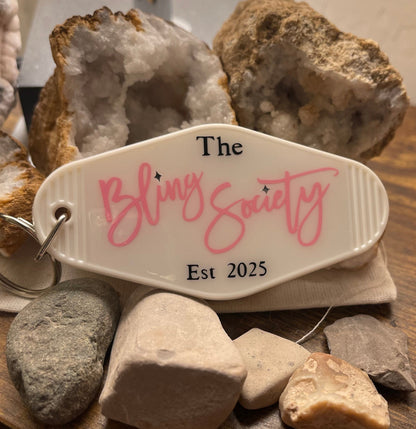Keychain with 'The Bling Society' logo on a wooden surface with rocks